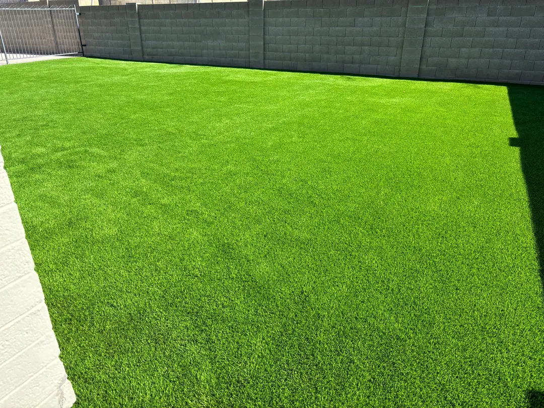 A bright, green artificial grass lawn bordered by a beige brick wall with a metal gate in one corner. The grass is evenly cut and well maintained, with shadows cast on one side.